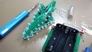 A picture of a table full of parts. Parts of the Christmas tree soldering kit, LED lights, a soldering iron and some batteries.